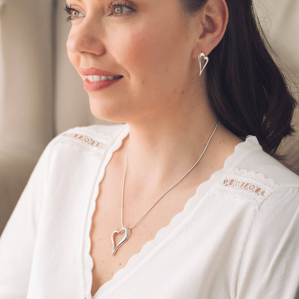 A woman wearing a delicate silver heart pendant necklace and matching heart stud earring, dressed in a white lace-trimmed top.