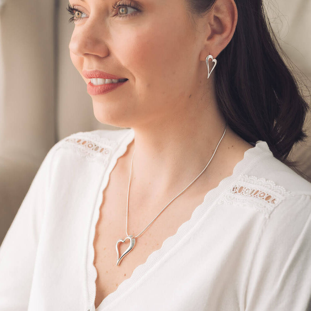 A woman wearing a delicate silver heart pendant necklace and matching heart stud earring, dressed in a white lace-trimmed top.