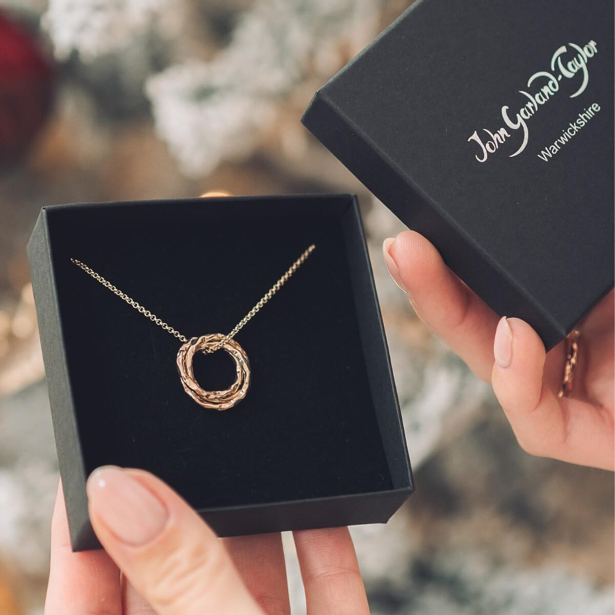 Rose gold necklace in a black box with 'John Hardy' branding, held by a hand against a blurred natural background.